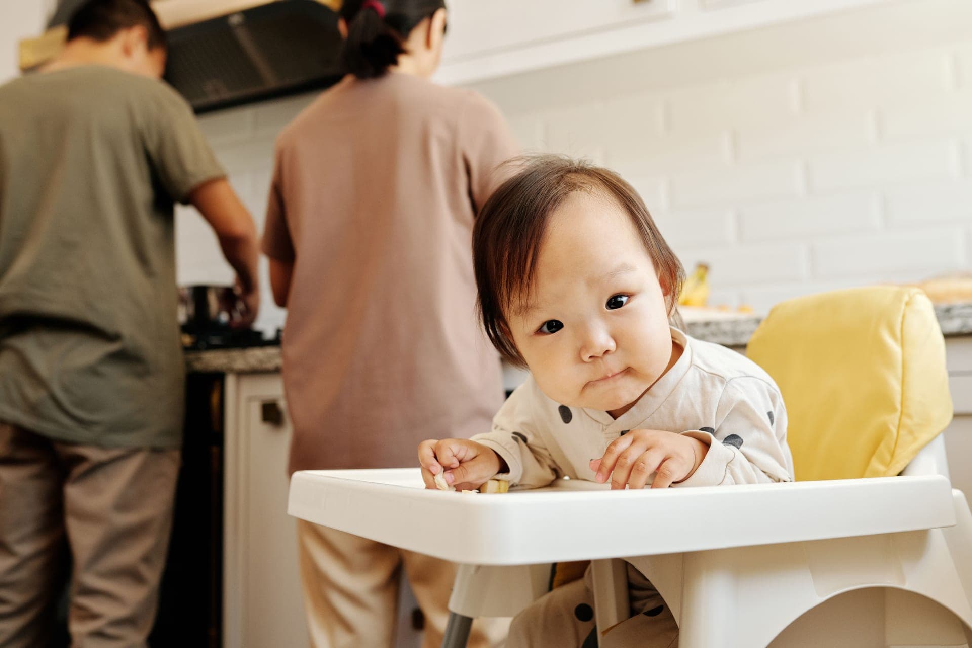A toddler sitting in a high chair while a parent prepares food in the kitchen (illustrative photo)