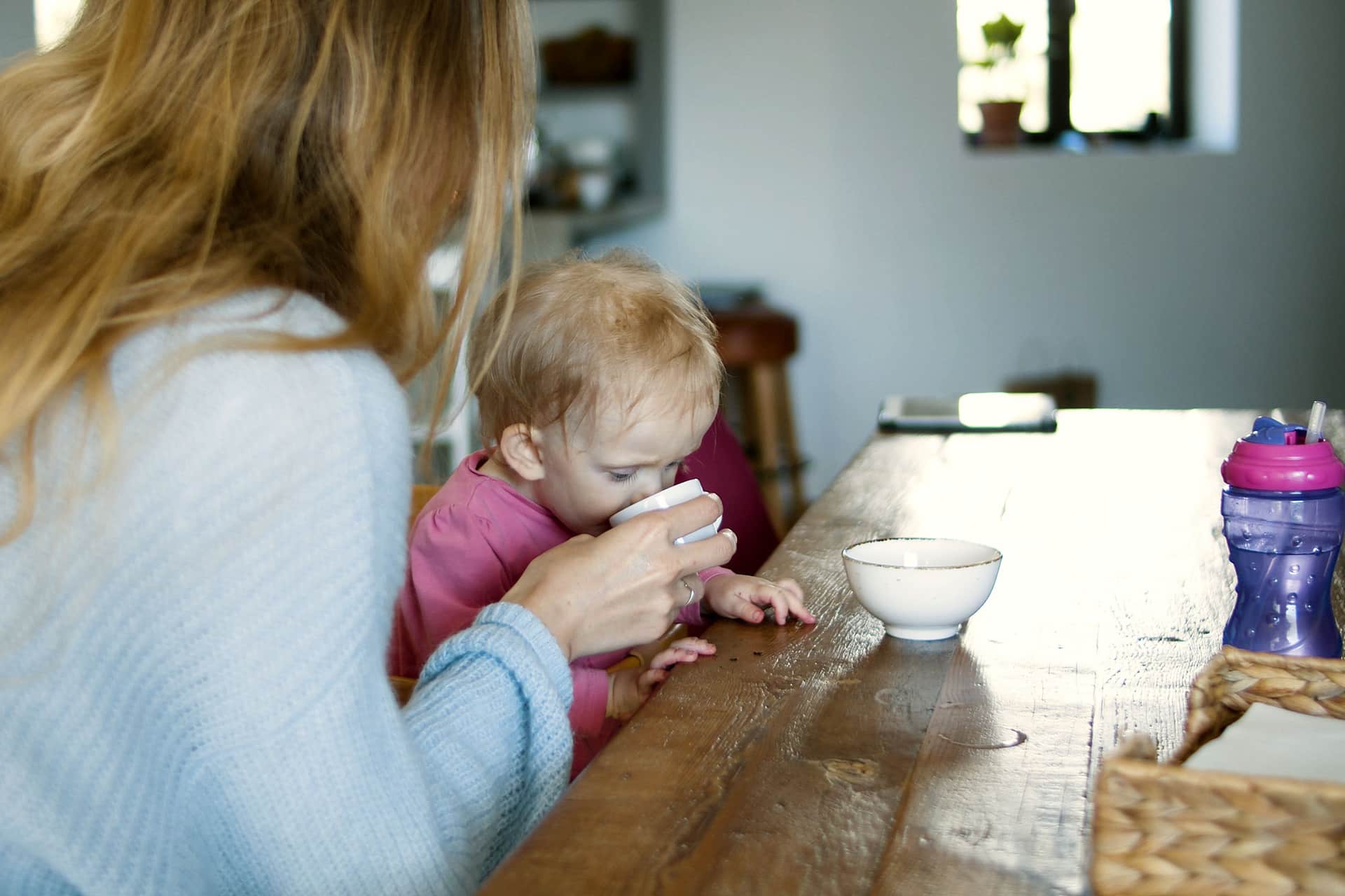 A parent helping their baby drink at a wooden table in a home kitchen (illustrative photo)