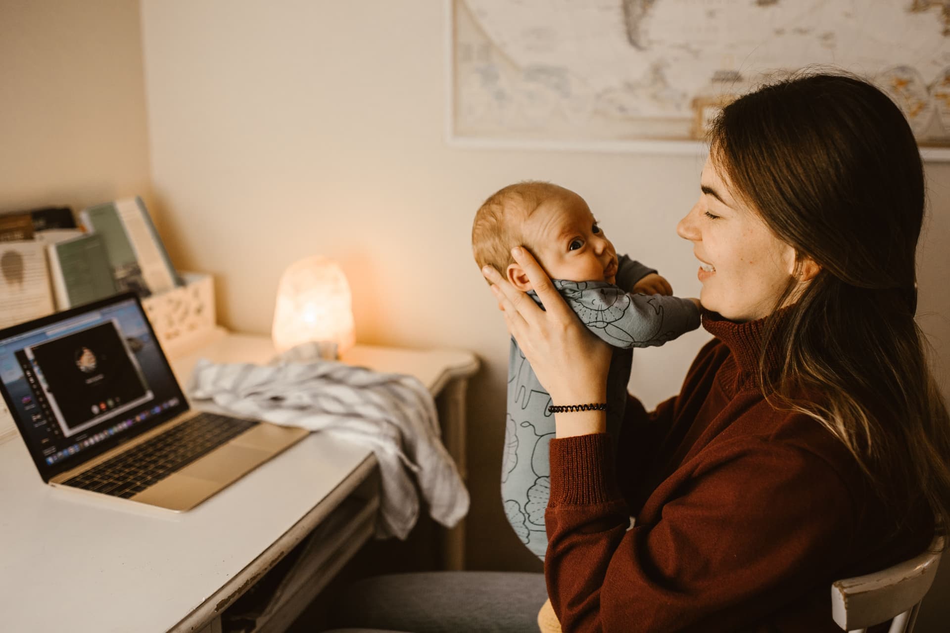 A parent holding their baby while working at a laptop in a home office (illustrative photo)